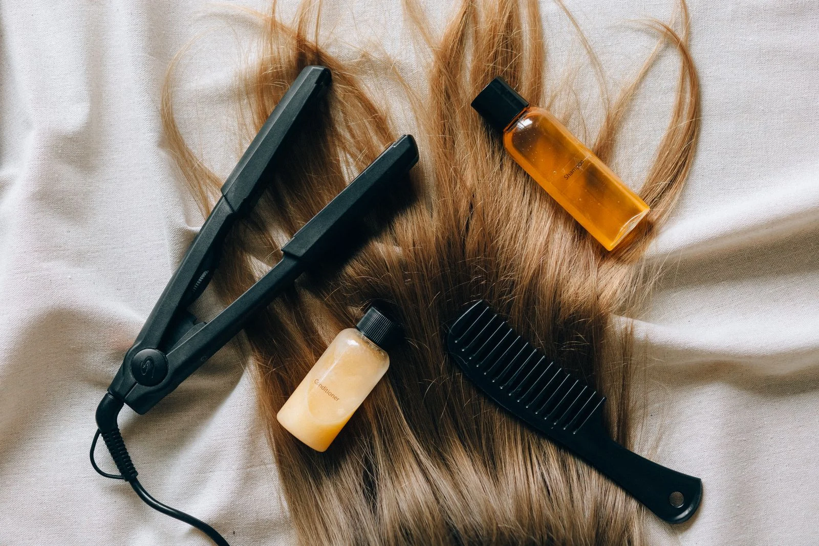Black hair straightener, wide-tooth comb and bottles of hair oil laid over long light-brown hair