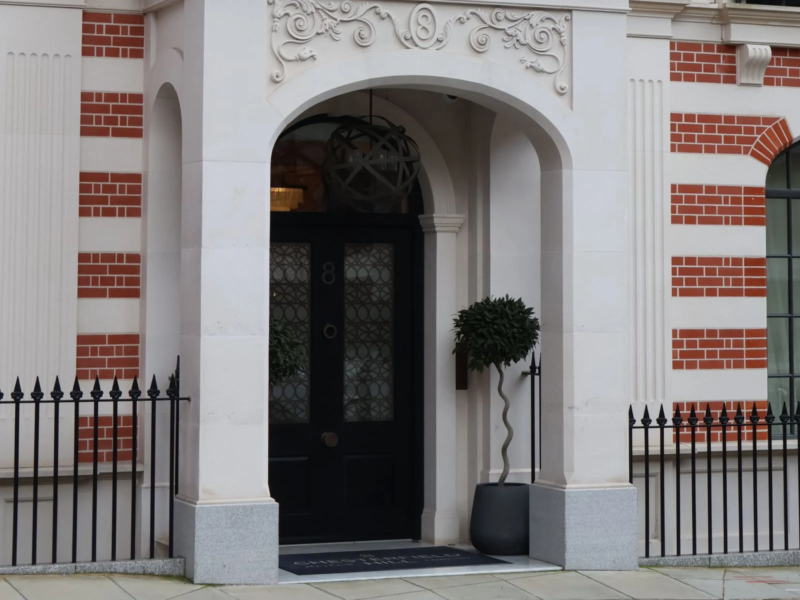 Arched stone doorway of a red-brick Mayfair townhouse with a black panelled door, topiary and iron railings