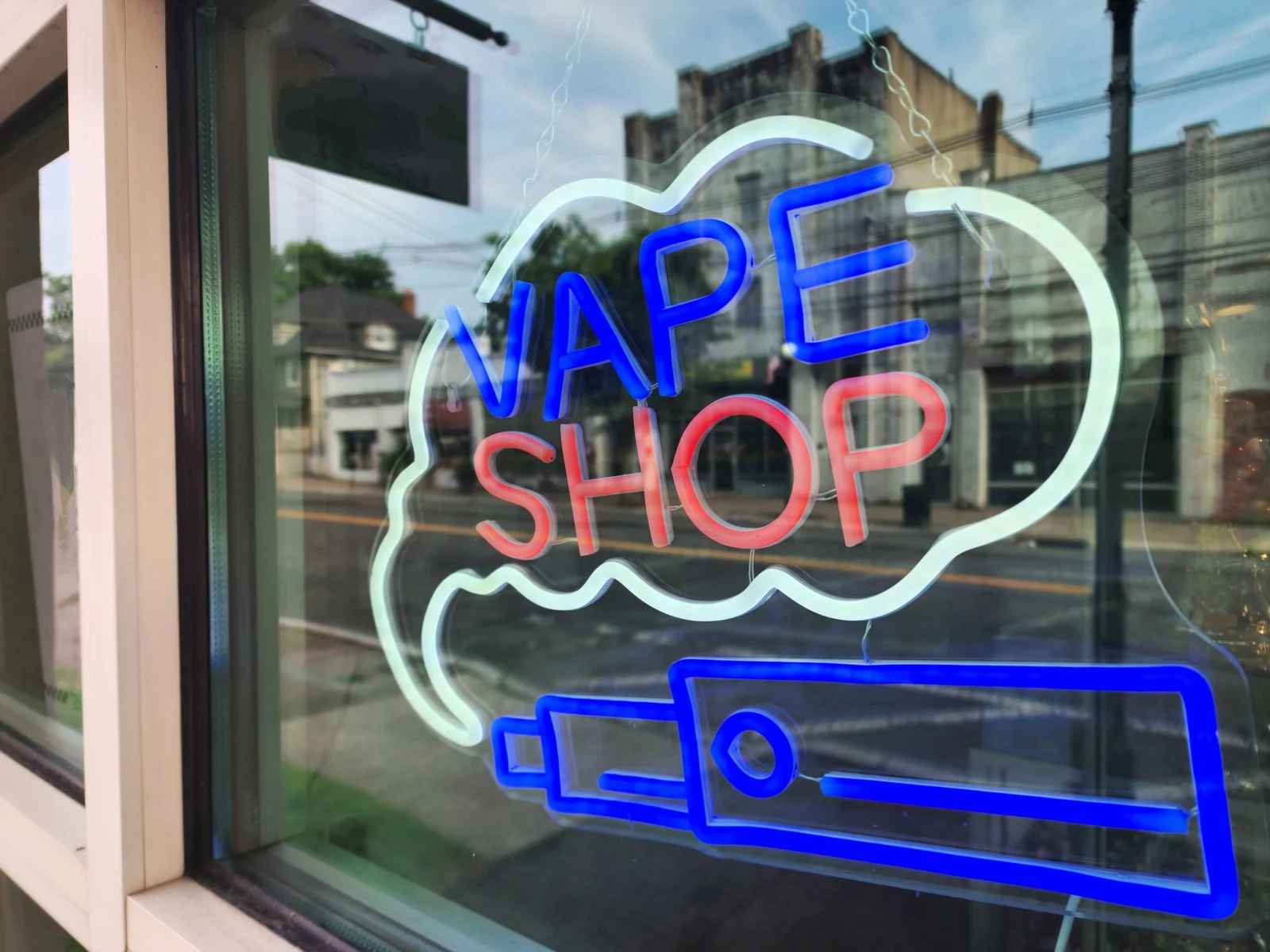 Neon vape shop sign glowing in a shop window, with the street reflected in the glass