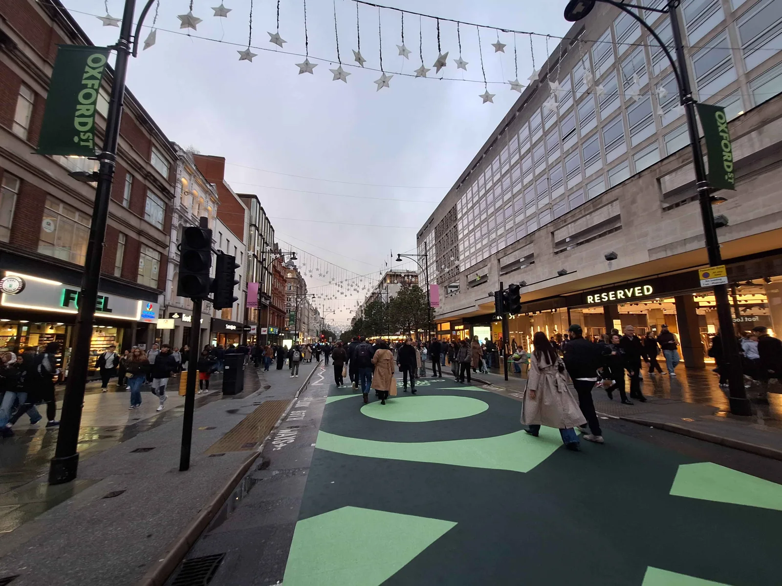 Shoppers walking down the middle of pedestrianised Oxford Street on a green painted carriageway under star lights